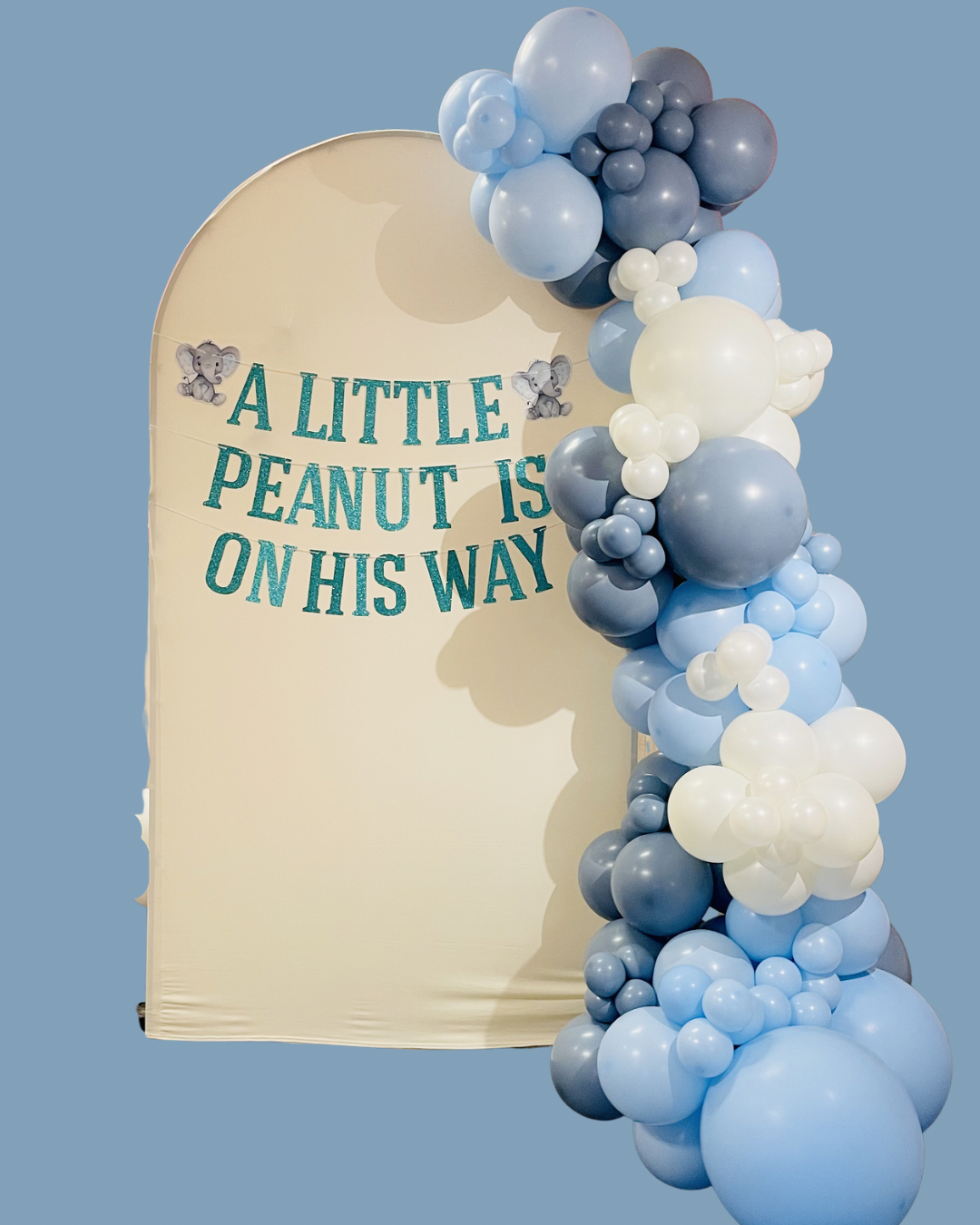 7.5ft cream fabric arch backdrop with banner “ A Little Peanut Is On His Way” and baby elephants. Coordinating baby blue, dusk blue and cream white organic balloon garland draped on the right side. 
